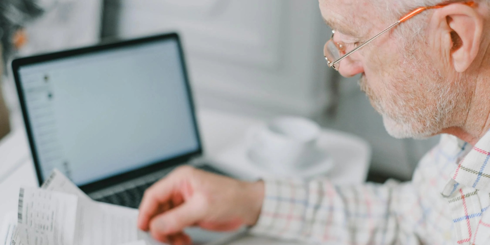 Homme senior examinant des documents médicaux avec un ordinateur portable à ses côtés.